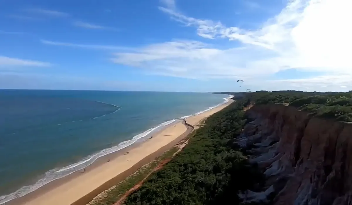 Vista aérea panorâmica de voo de parapente Arraial sobre a praia, falésias coloridas e mar azul turquesa.