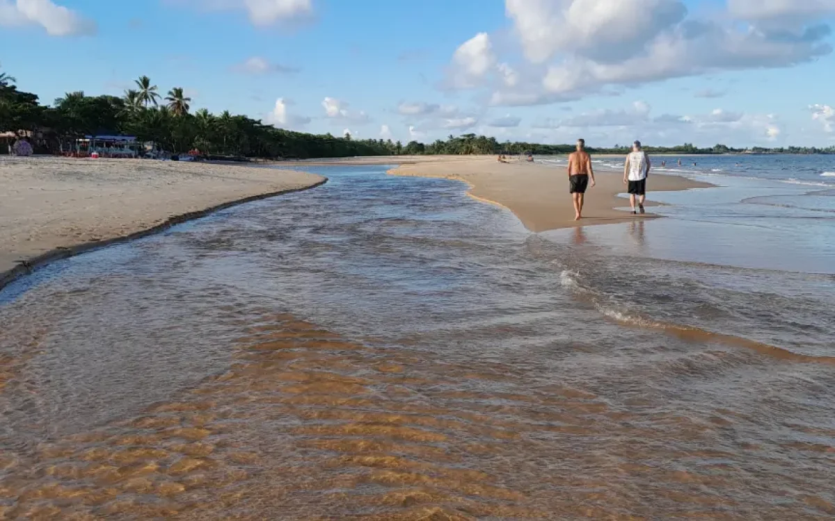 Riacho Barramares encontra o mar na Praia de Taperapuã, Porto Seguro, com dois homens caminhando na areia dourada sob um céu azul.