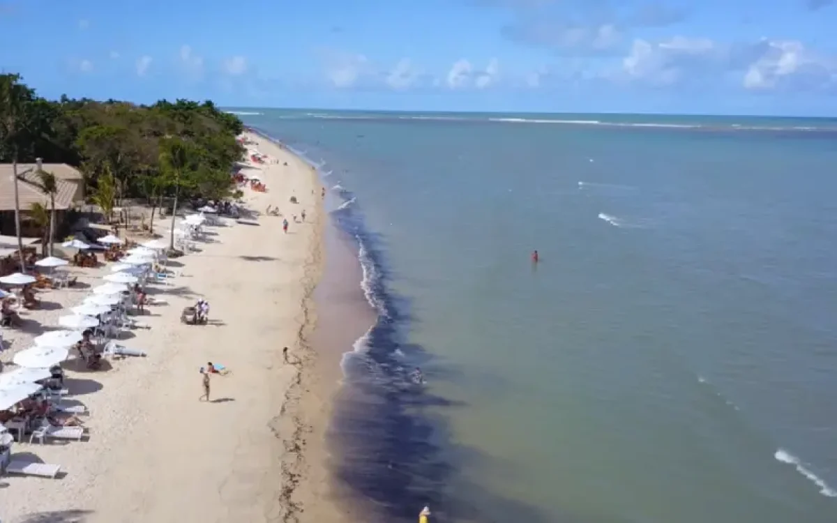 Vista aérea da Praia do Mutá em Porto Seguro, com faixa de areia clara, coqueiros, guarda-sóis e mar calmo em tons de azul e verde.