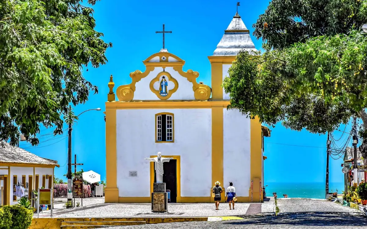 Igreja Nossa Senhora d'Ajuda em Arraial d'Ajuda, Bahia, com vista para o mar e árvores verdes.