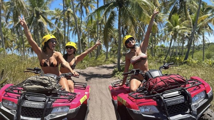 Três mulheres sorridentes usando capacetes amarelos em dois quadriciclos Honda vermelhos em uma trilha de areia cercada por coqueiros em Trancoso, Bahia.