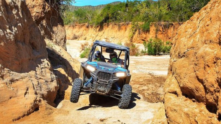 Um UTV azul e preto, com duas pessoas usando capacetes e bandanas faciais verdes, navegando por um cânion estreito de terra vermelha com vegetação de Caatinga no fundo em Trancoso, Bahia. Um pneu dianteiro está no ar.
