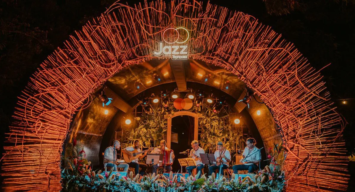 Palco rústico e iluminado do Festival de Jazz em Trancoso SEO, apresentando seis músicos tocando instrumentos de corda e sopro sob um arco de madeira trançada em Porto Seguro.