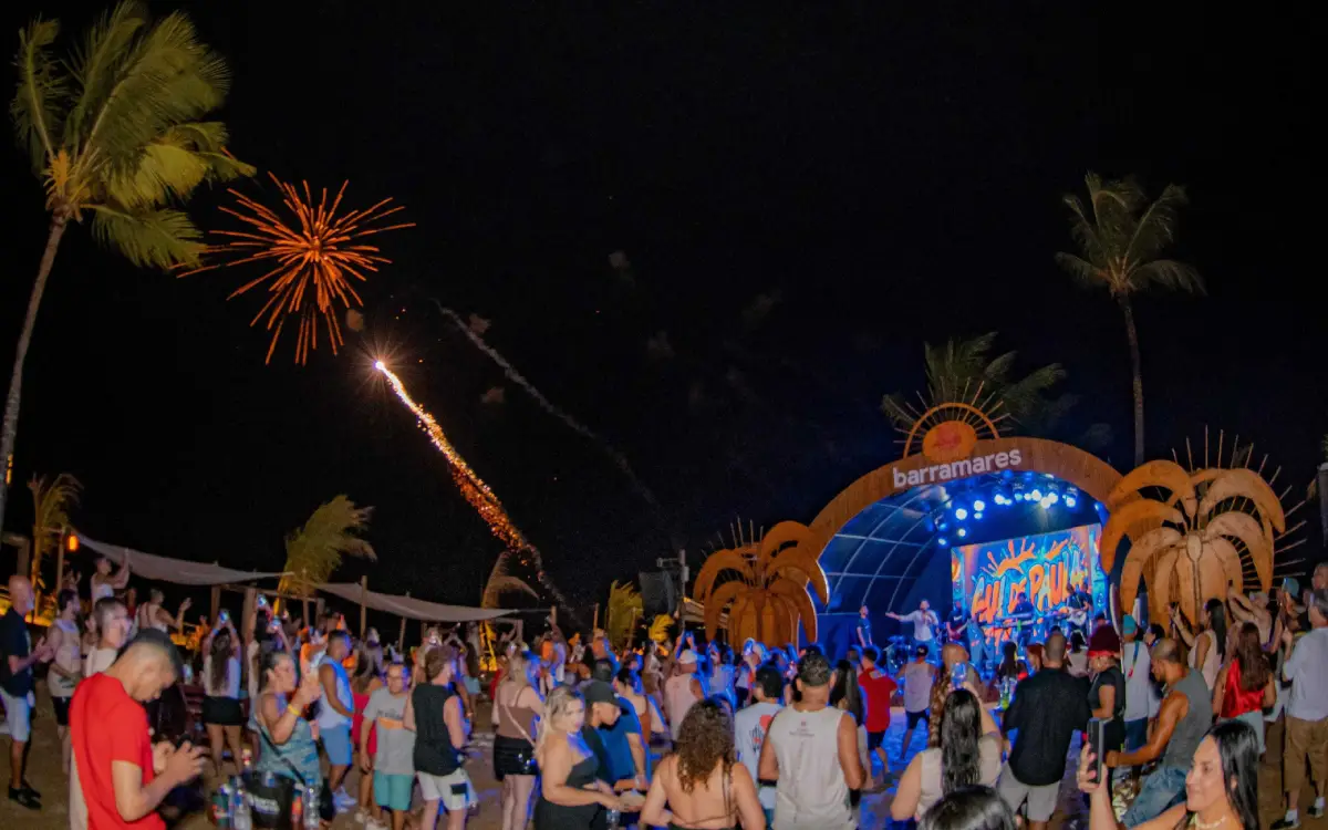 Pessoas dançando em um luau noturno na praia do Barramares em Porto Seguro, com palco iluminado, palmeiras e fogos de artifício no céu.