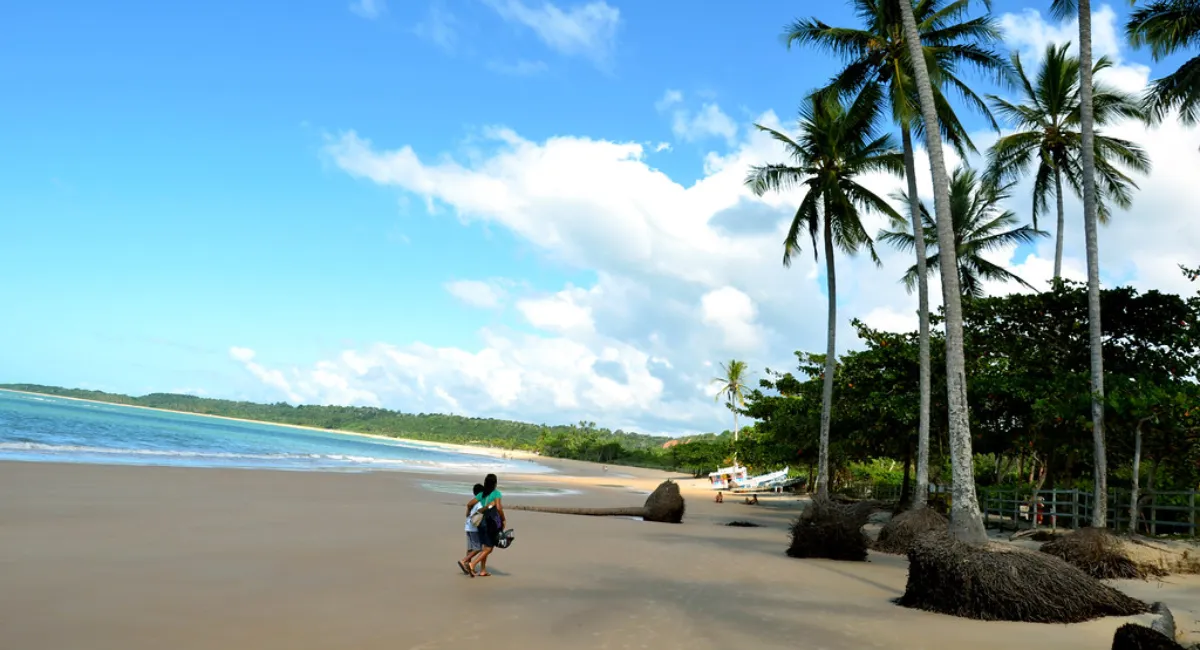Praia dos Coqueiros em Trancoso, Bahia, com coqueiros e duas pessoas caminhando na areia.