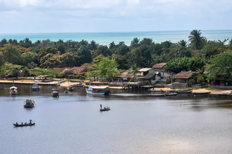 Vista panorâmica aérea da vila rústica de Caraíva, na Bahia, mostrando o encontro do rio Caraíva com o oceano Atlântico. Barcos coloridos estão ancorados no rio calmo e canoas cruzam as águas. A orla é arborizada com vegetação nativa densa e casinhas de pescadores estão dispostas ao longo da margem. O céu está claro e o mar ao fundo tem tons de turquesa.