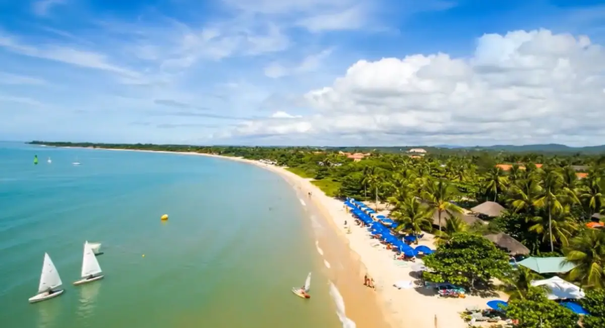 Vista aérea da Praia do Mutá em Porto Seguro, Bahia, mostrando a faixa de areia clara, guarda-sóis azuis, barcos a vela no mar verde esmeralda e muitos coqueiros.